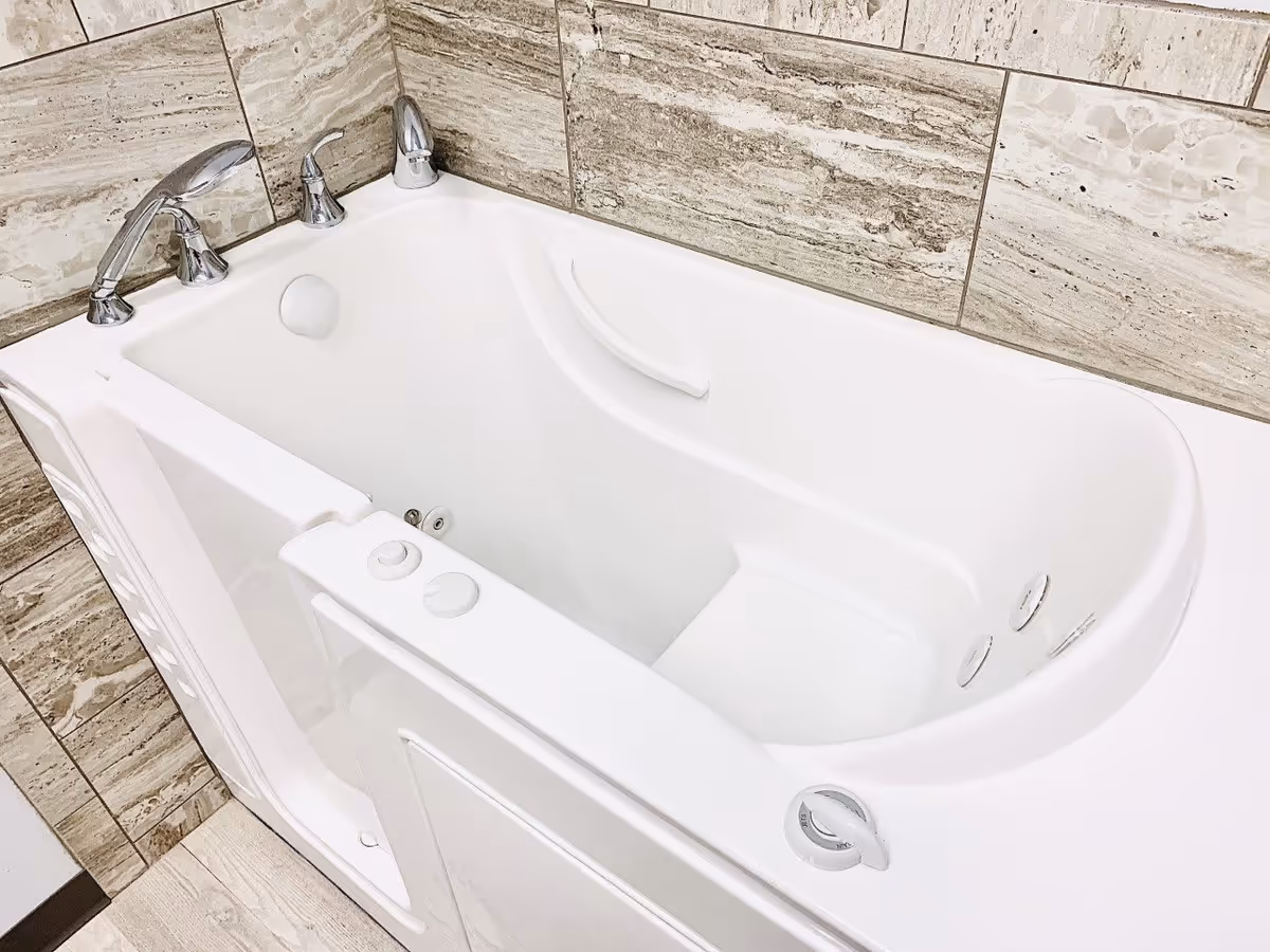 A white walk-in bathtub with chrome faucet handles and a handheld showerhead, surrounded by beige and brown marble tiles on the walls.