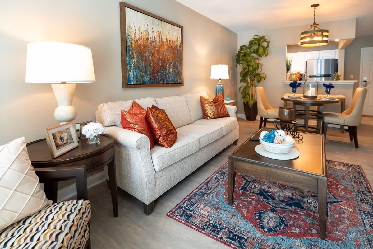 A cozy living room area featuring a beige sofa with red and orange decorative pillows, a wooden coffee table with a decorative bowl, a patterned rug, a side table with a lamp and framed photo, a large plant in the corner, and a dining table with four chairs in the background near a kitchen area.