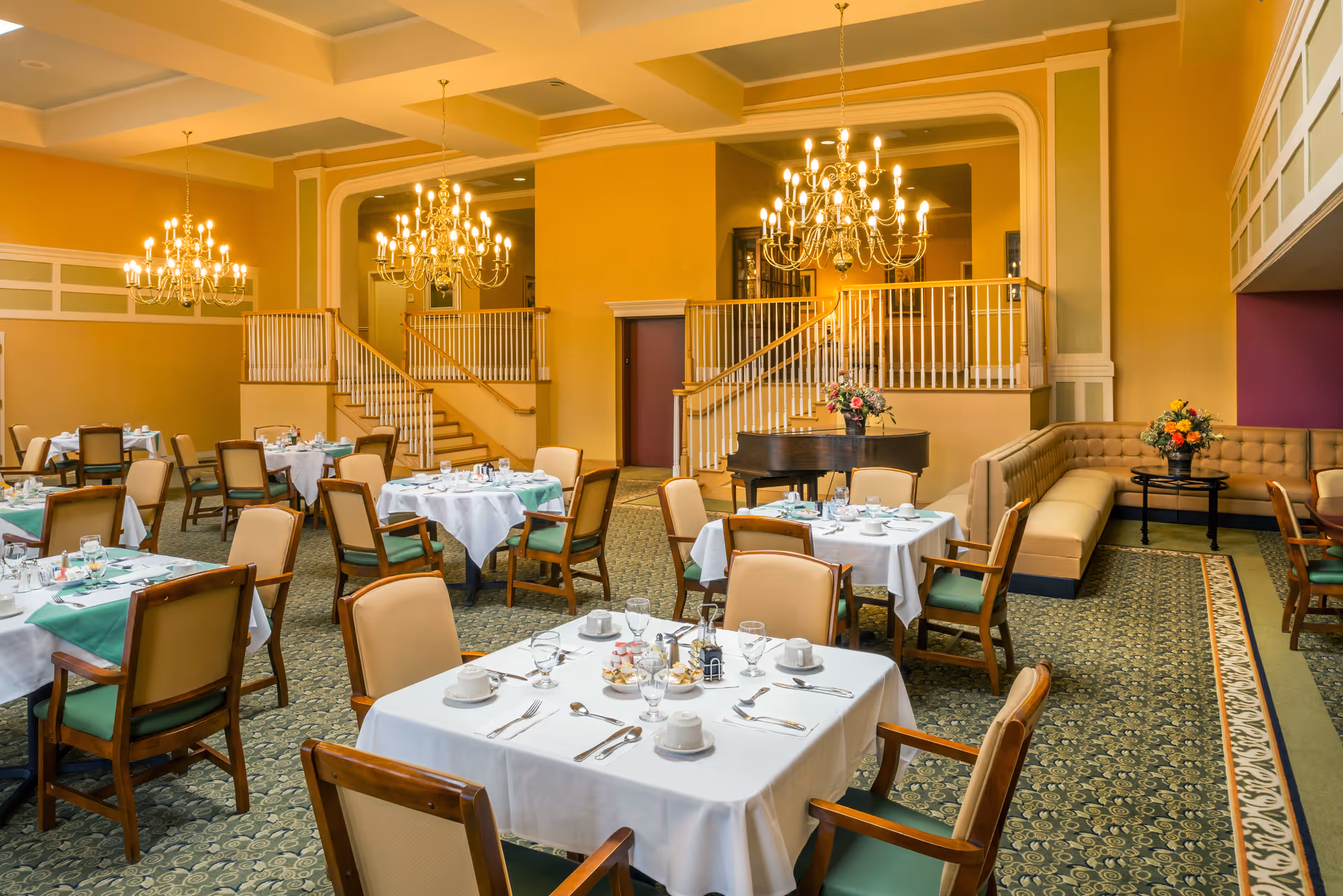 A spacious dining room with multiple tables covered in white tablecloths and set with cups, glasses, and silverware. The room features elegant chandeliers, a grand piano with a flower arrangement, and a staircase with white railings. There is a long beige cushioned bench along one wall with a small table holding a flower arrangement.