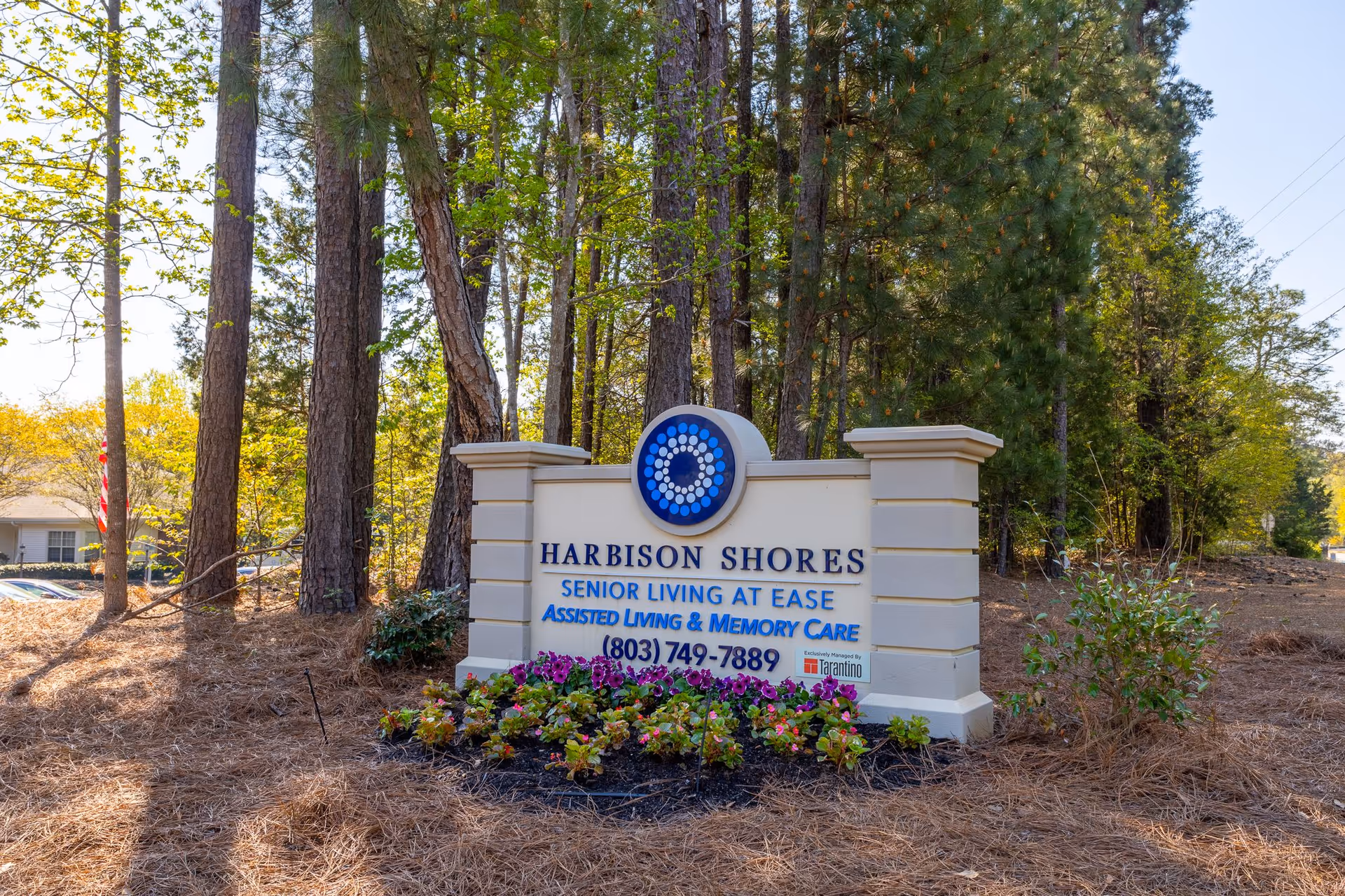 Outdoor view of a sign for Harbison Shores Senior Living at Ease, Assisted Living & Memory Care, surrounded by trees and flowers with a clear sky in the background.