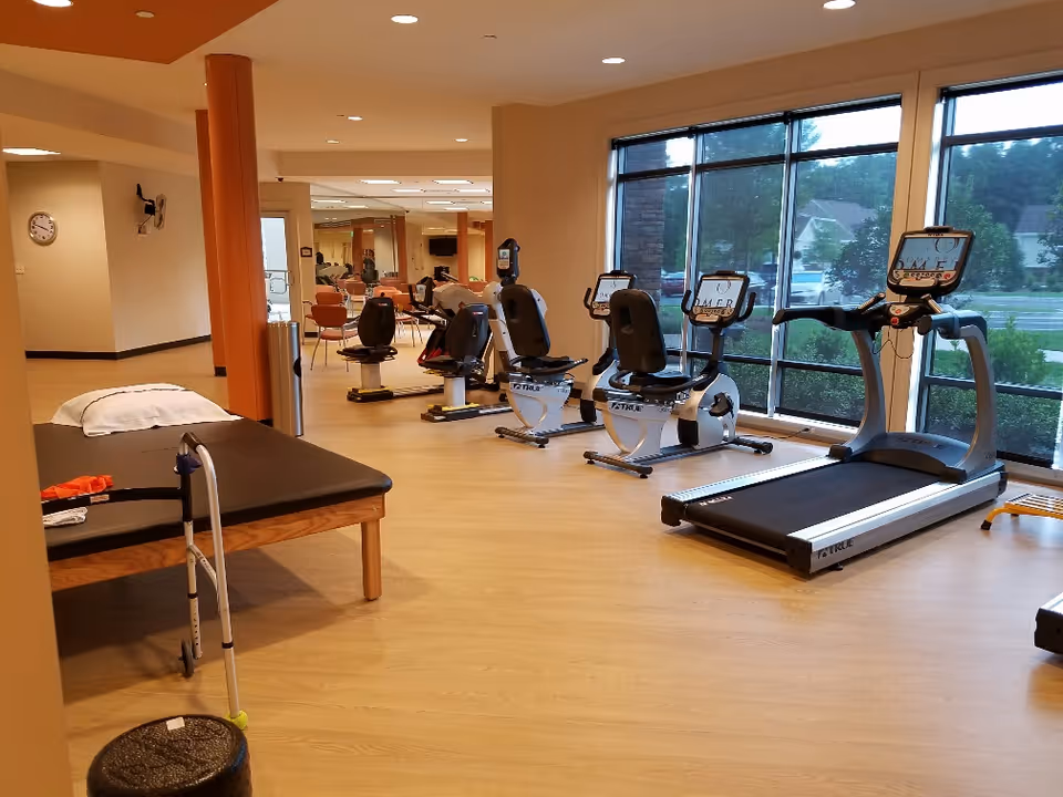 A fitness room with exercise equipment including a treadmill and several stationary bikes facing large windows. There is a padded exercise table with a pillow and a walker nearby. The room has wooden flooring, beige walls, and large mirrors on one side reflecting the equipment and chairs.