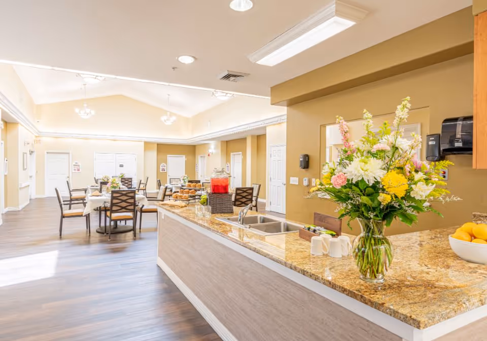 Bright dining area with a granite service counter and a vase of flowers in the foreground and tables and chairs in the background.