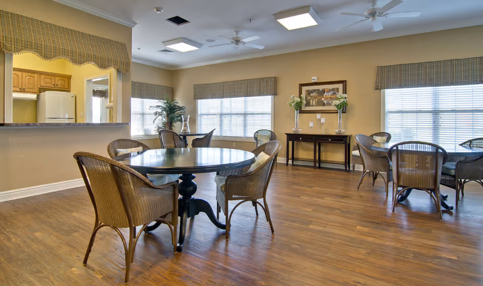 A bright dining room in a senior living facility with round tables and wicker chairs arranged on a wooden floor. Large windows with plaid valances let in natural light. A side table with decorative vases and flowers is against the wall, and a kitchen area is visible through a pass-through window.