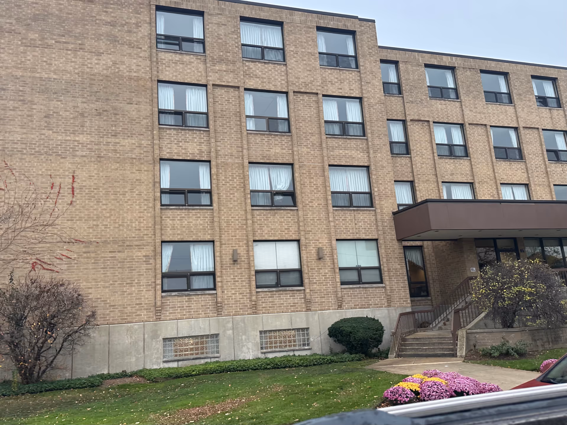 Exterior view of a multi-story brick building with multiple windows, a small staircase leading to an entrance with a brown canopy, some bushes, a tree with red leaves, and a flower bed with pink and yellow flowers in front.