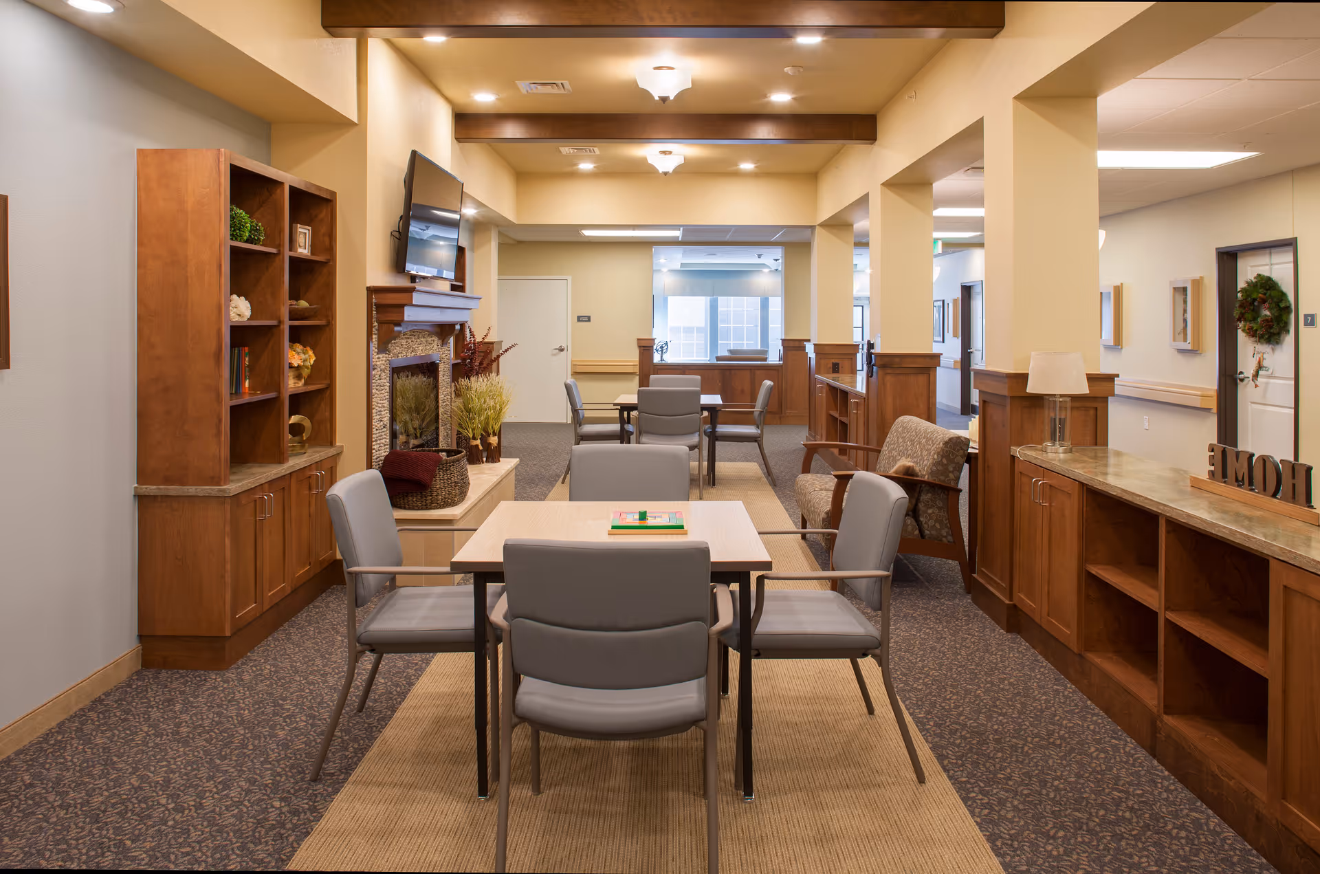 A cozy common area in a senior living facility featuring a table with four gray chairs, a fireplace with decorative plants, a wall-mounted TV above the fireplace, wooden cabinetry and shelving, and additional seating with an armchair. The space is warmly lit with ceiling lights and has a carpeted floor with a beige rug under the table.