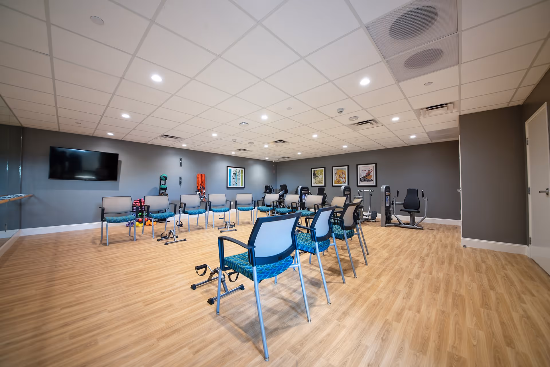 A spacious exercise room with wooden flooring and gray walls, equipped with chairs arranged in a semi-circle, various exercise equipment including stationary bikes and hand weights, a wall-mounted TV, and framed artwork on the walls.