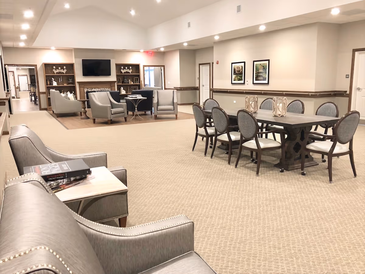 Spacious senior living common room with multiple seating areas, a TV above a fireplace and a long dining table with chairs.