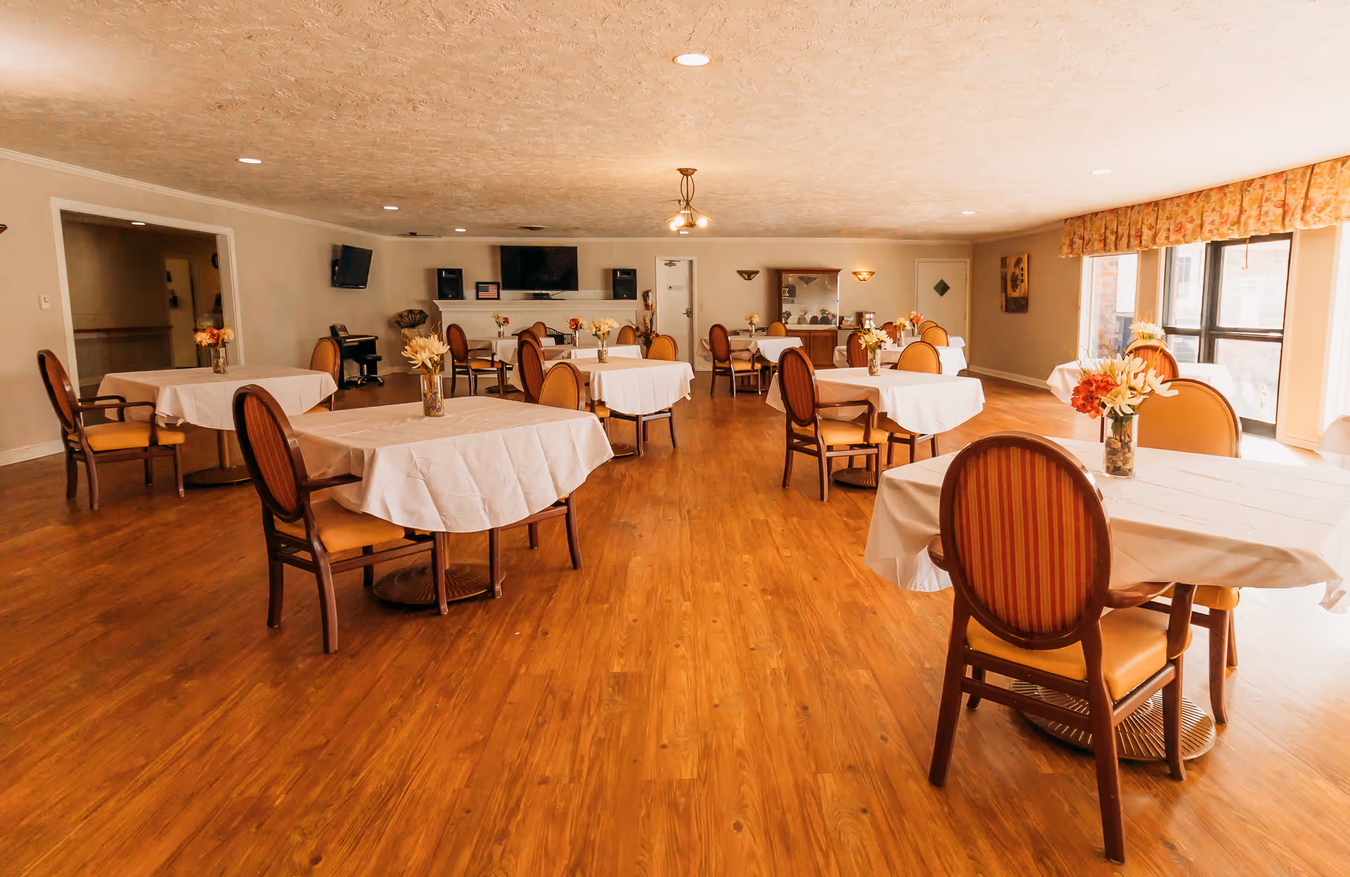 Spacious dining room with round tables covered in white tablecloths, wooden chairs, and floral centerpieces.