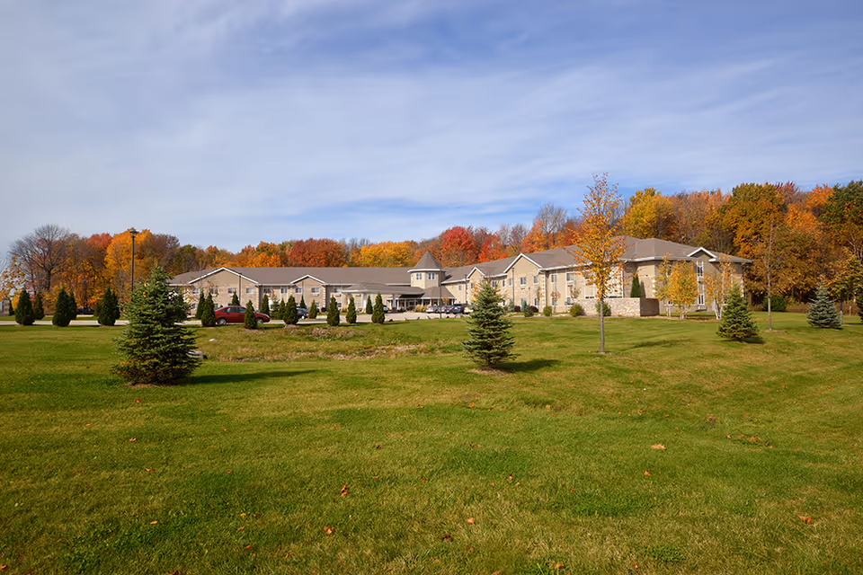 Wide view of a large, single-story retirement community building with a stone facade surrounded by a well-maintained green lawn and small trees. The background features a line of trees with autumn foliage under a partly cloudy sky.
