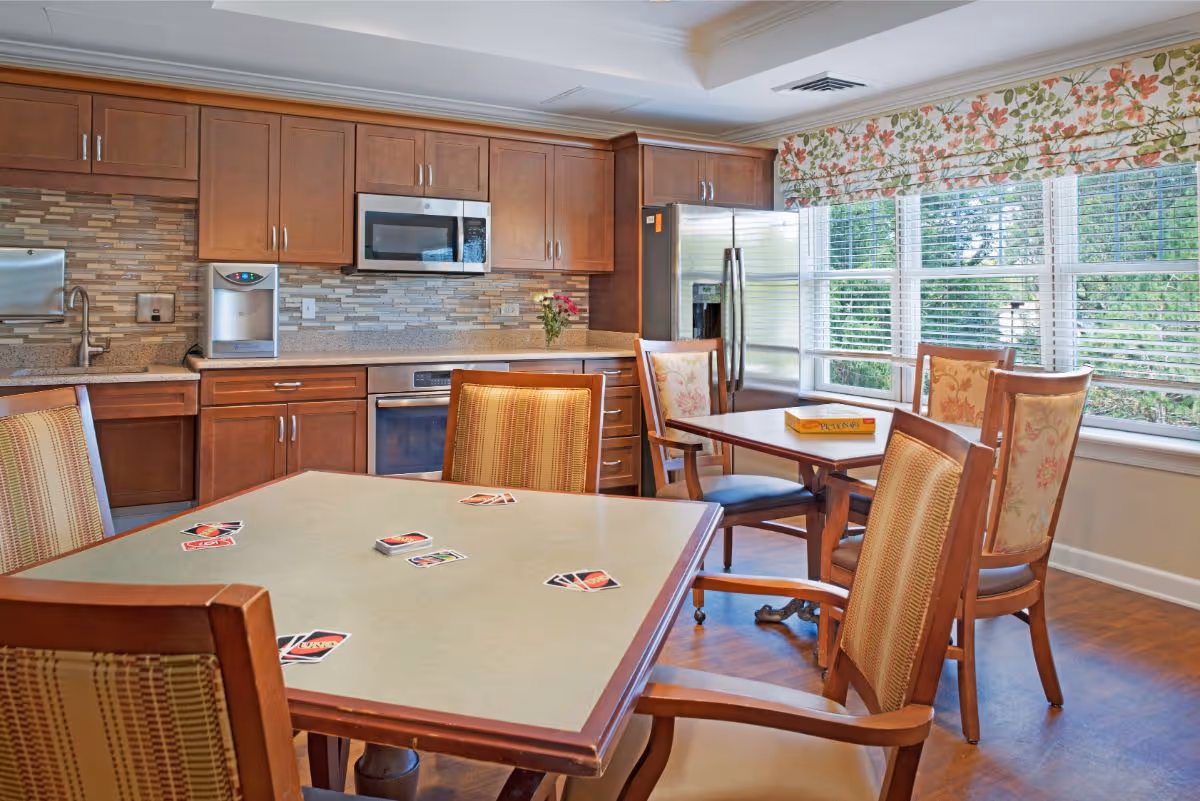 Sunlit communal dining/activity room with tables and chairs, playing cards on a table, wooden cabinetry and stainless steel appliances along the back wall.