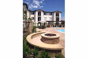 Courtyard with a circular stone fire pit seating area and pool in front of a three-story residential building under a blue sky.