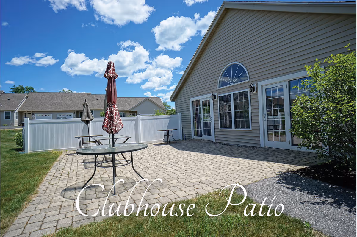 Outdoor patio area with stone pavers, glass tables with closed umbrellas, adjacent to a building with large windows and a door under a blue sky with scattered clouds.