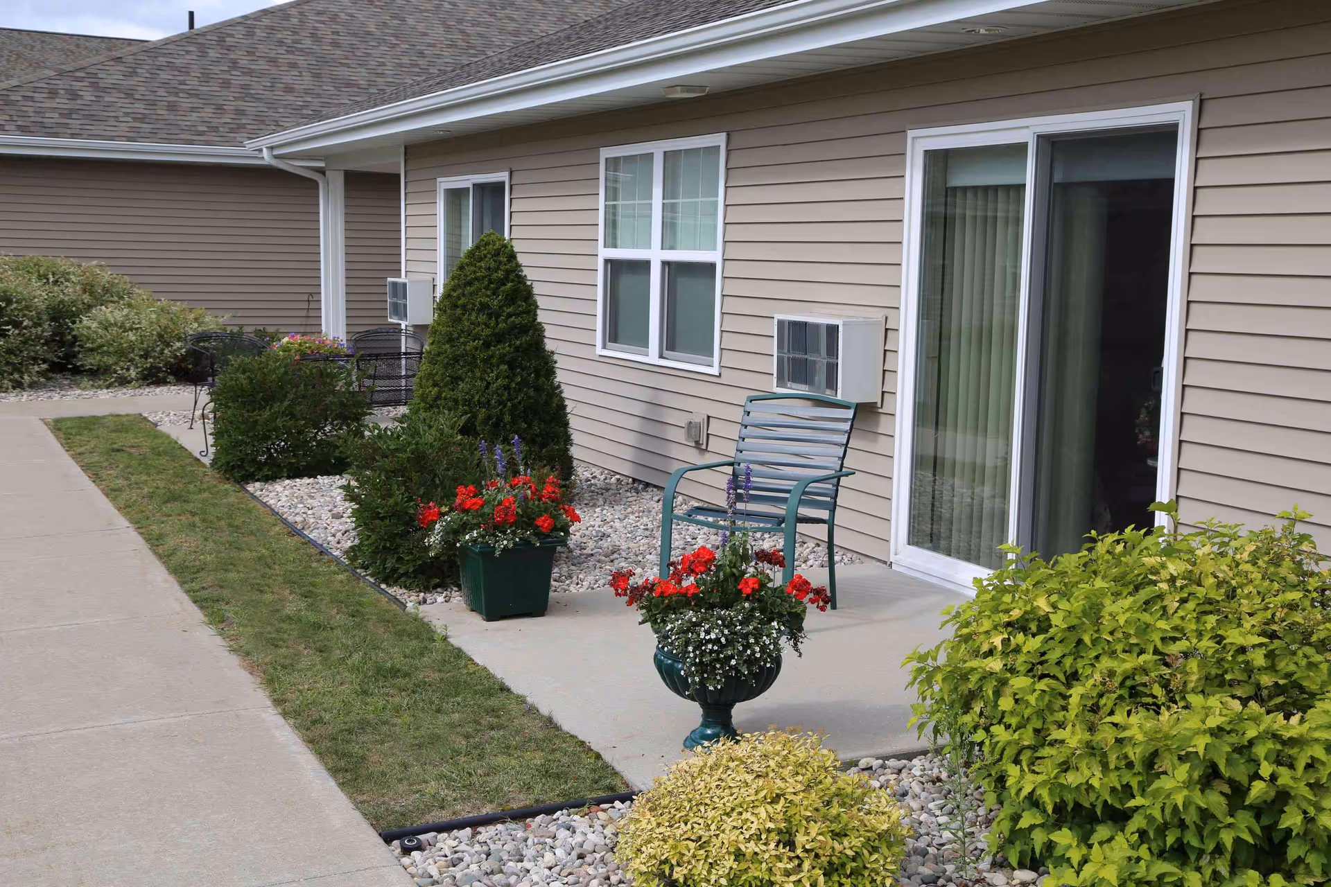 Outdoor patio area of a senior living facility with beige siding, a sliding glass door, a window with an air conditioning unit, a metal chair, and several planters with red and white flowers surrounded by bushes and landscaping rocks.