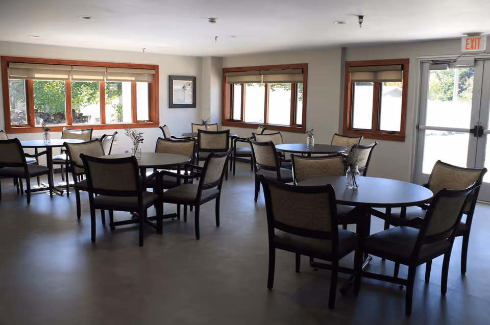 Well-lit dining room with round tables and upholstered chairs arranged near windows and an exit door.