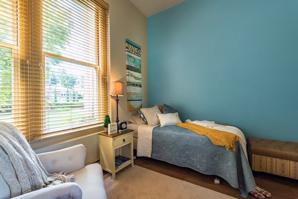 A cozy bedroom with a single bed covered in a blue patterned bedspread and a mustard yellow throw blanket. There are several pillows on the bed. Next to the bed is a small cream-colored nightstand with a lamp, a small plant, a clock, and a decorative sign that says 'I love MOM'. A comfortable armchair with a knitted throw blanket is positioned near a large window with wooden blinds, letting in natural light. The walls are painted teal and cream, and there is a colorful abstract painting hanging above the bed. A cushioned bench is placed at the foot of the bed.