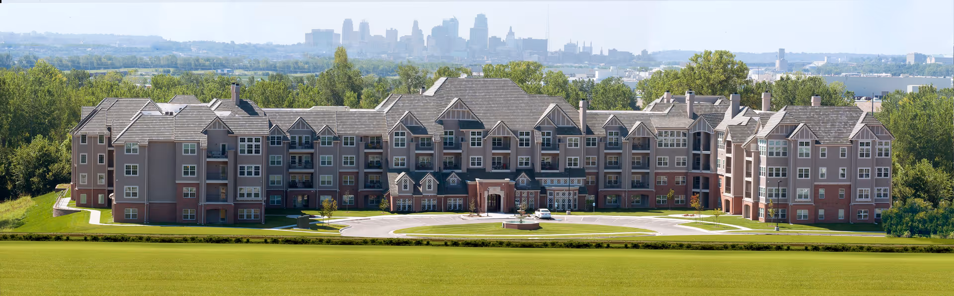 Front exterior of a large multi-story senior living building set on a landscaped lawn with a distant city skyline behind it.