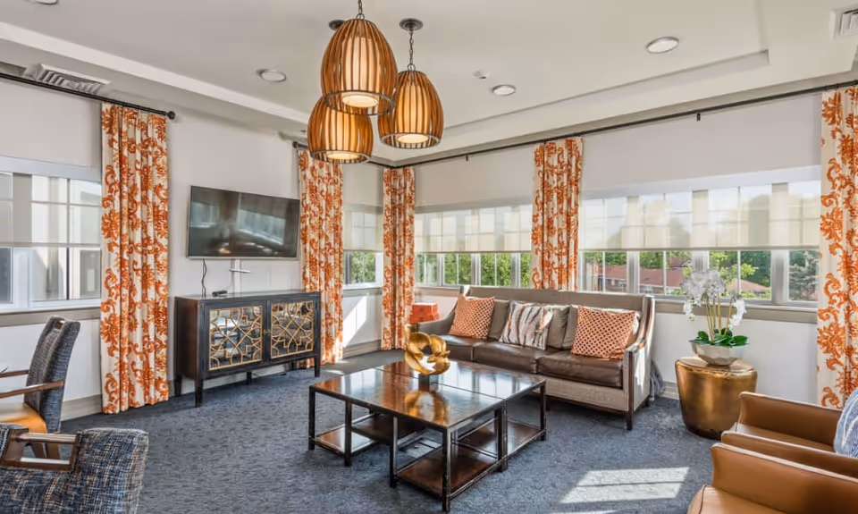 A bright and cozy living room with large windows covered by white roller shades and orange patterned curtains. The room features a brown leather sofa with decorative pillows, a dark wooden coffee table with a gold decorative piece, two armchairs, a TV mounted on the wall above a stylish cabinet, and three pendant lights hanging from the ceiling. A small round table with a white orchid plant is placed near the sofa.