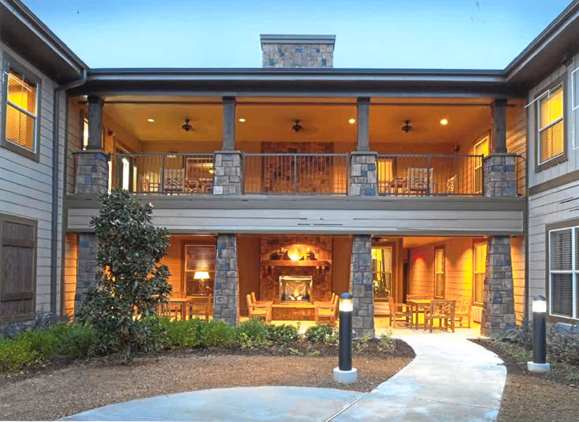 Exterior view of a two-story senior living facility building at dusk with a covered patio area on the ground floor featuring a lit fireplace and seating, and a balcony on the upper floor with additional seating. The building has stone pillars and warm lighting inside the rooms.