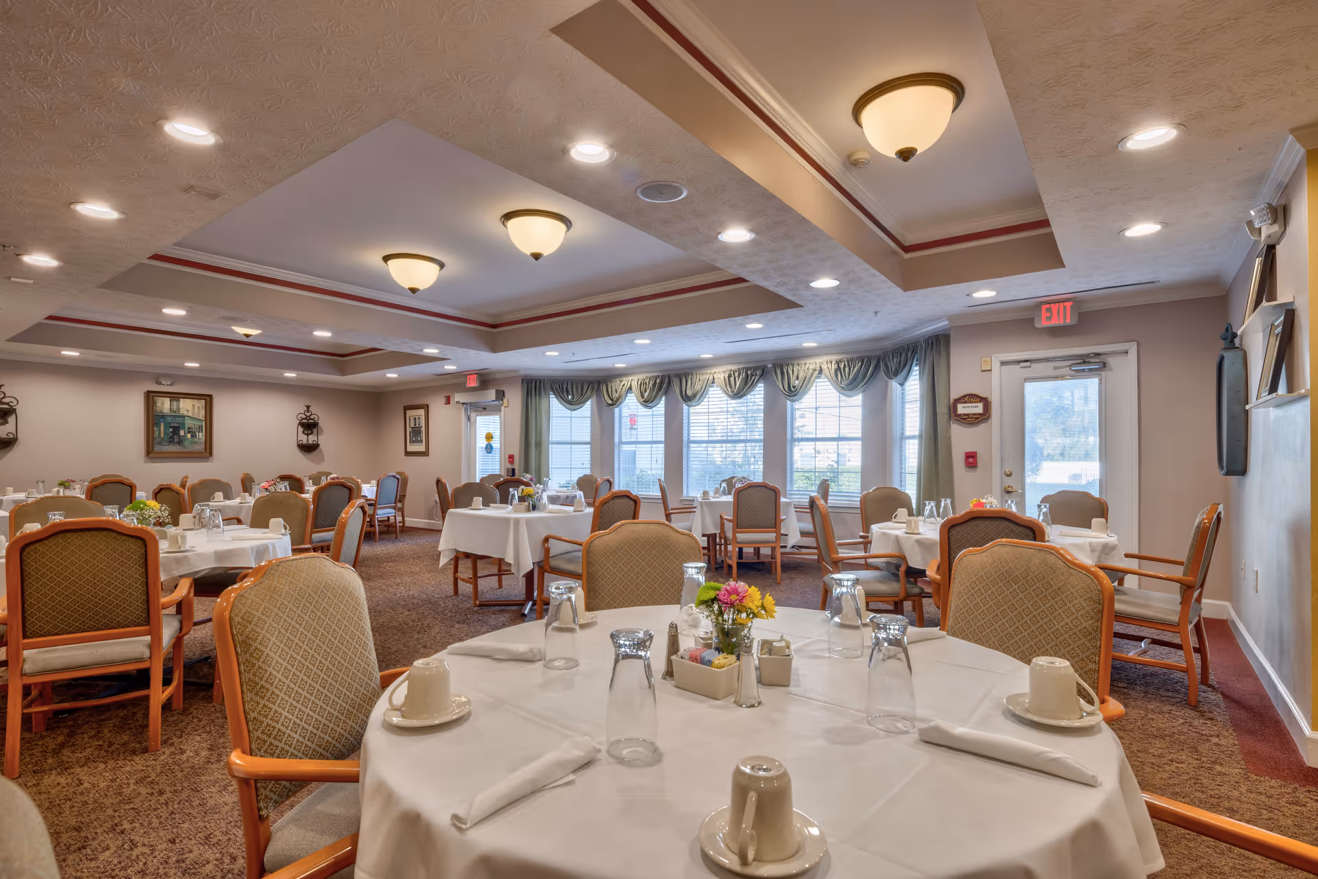 A spacious dining room with multiple round tables covered with white tablecloths, each set with cups, glasses, napkins, and small flower arrangements. The room has carpeted floors, beige walls, and large windows with green valances allowing natural light to enter. Ceiling lights and decorative wall art are visible, creating a warm and inviting atmosphere.