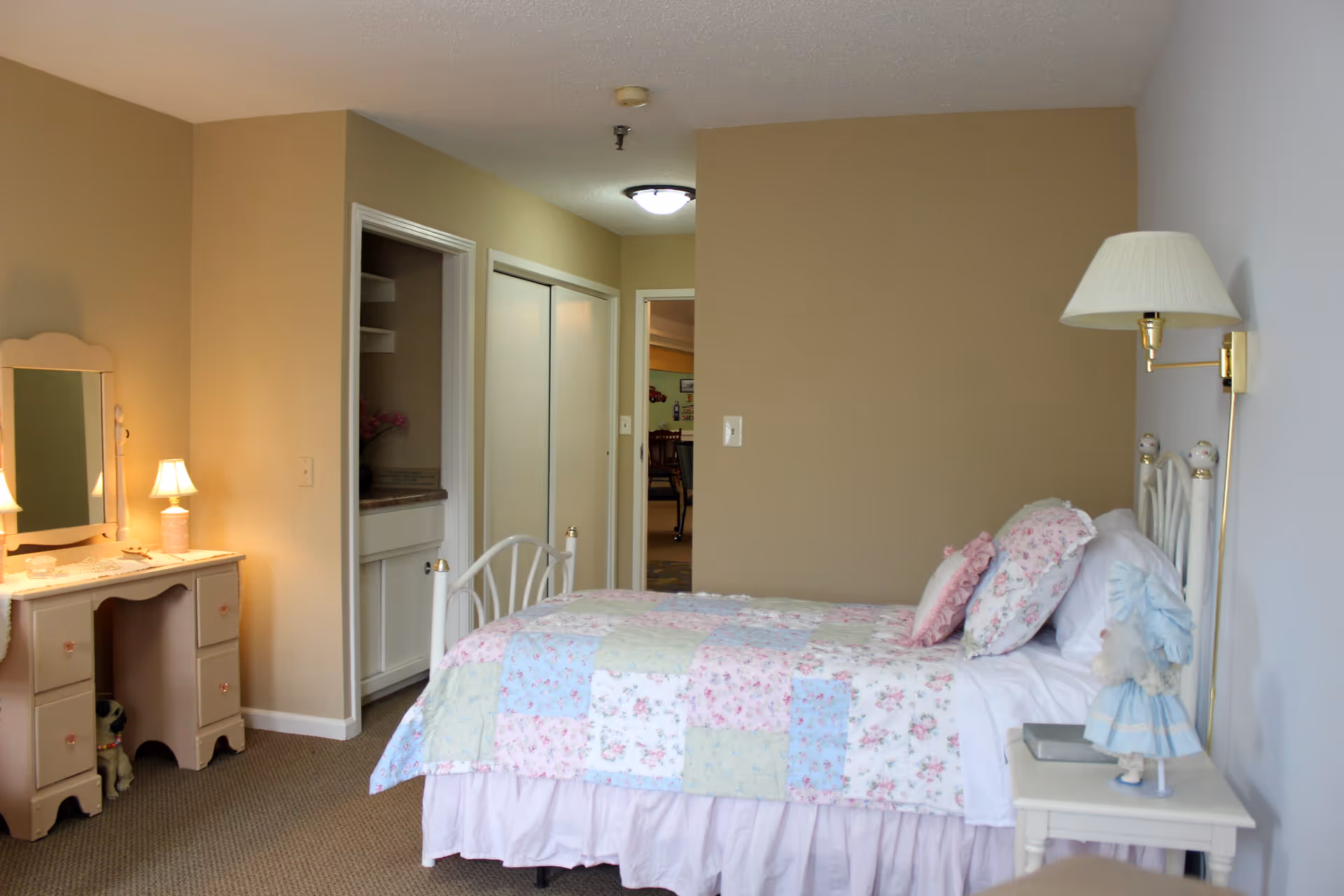 A cozy bedroom in a senior living facility featuring a single bed with a floral patchwork quilt and multiple pillows. To the right of the bed is a small white nightstand with a doll and a book, and a wall-mounted lamp above it. On the left side of the room is a vintage-style vanity with a mirror and a small lamp. The room has beige walls and carpeted flooring, with a closet and an open doorway leading to another room visible in the background.