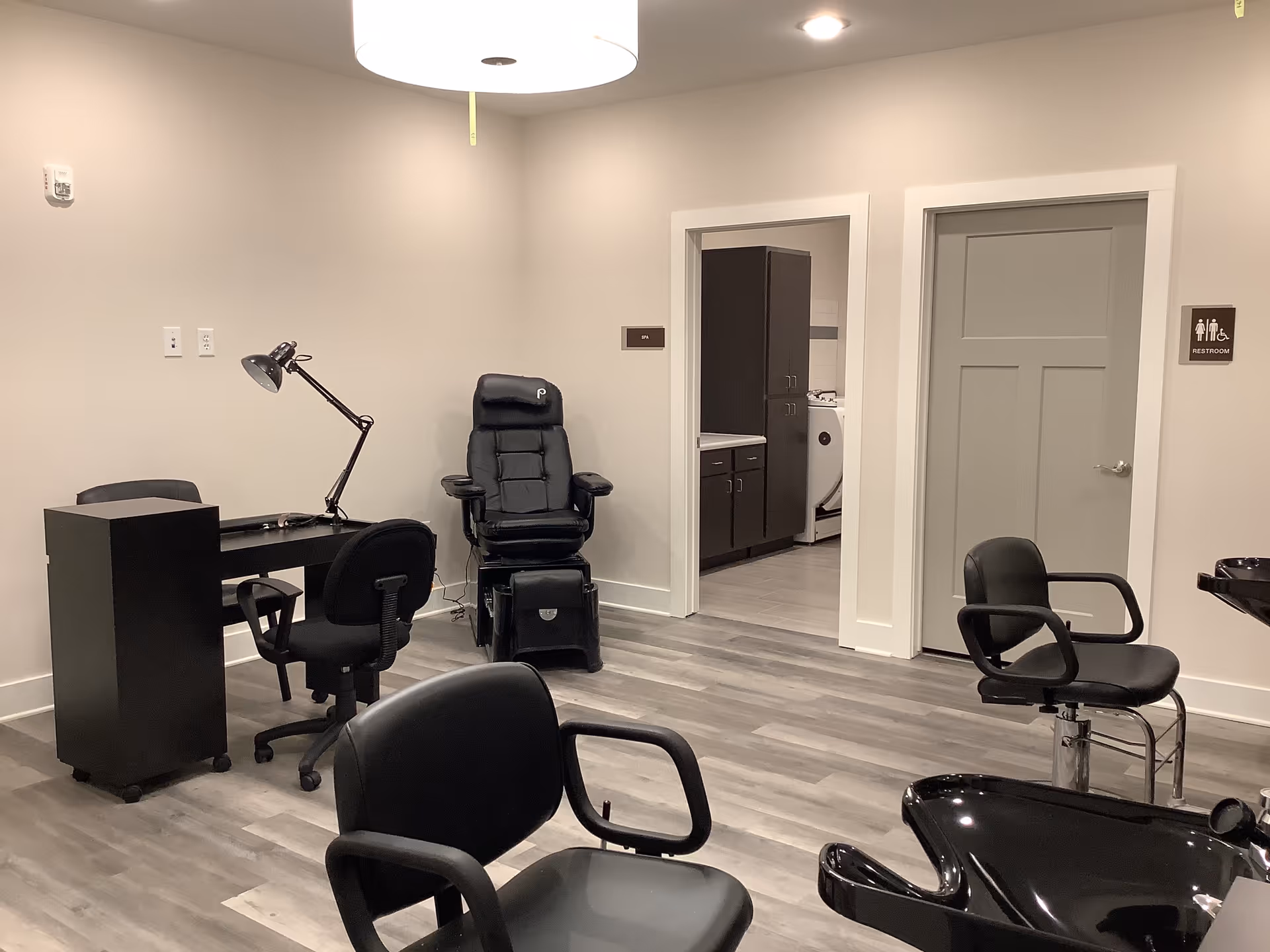 Interior view of a salon area in a senior living facility with black salon chairs, a manicure table with a lamp, a spa chair, and a door labeled restroom. The room has light-colored walls and wood flooring.