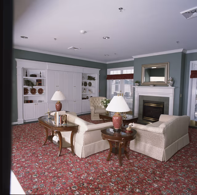 A cozy living room with beige upholstered sofas arranged around a wooden coffee table and side tables with lamps. The room features a red patterned carpet, green walls, a white fireplace with a mirror above it, and built-in white shelves displaying decorative items and books. Large windows with white blinds and red valances allow natural light into the space.
