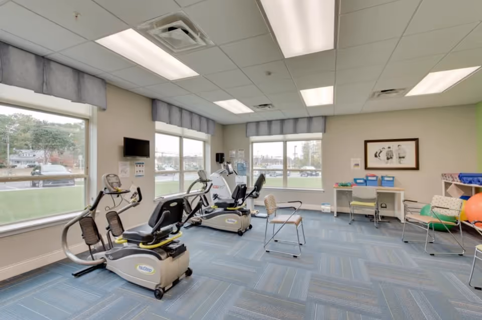A bright exercise room with large windows, two NuStep recumbent exercise bikes, several chairs, a desk with storage bins, and exercise balls on a rack. The room has blue carpet tiles and fluorescent ceiling lights.