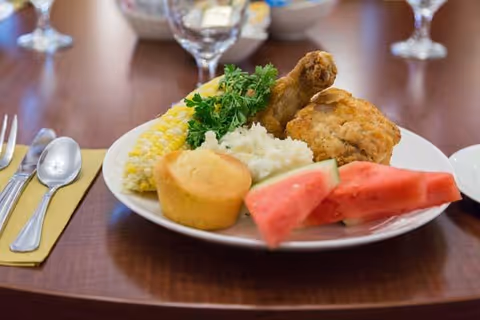 A plate of food on a wooden table featuring fried chicken, mashed potatoes with gravy, corn on the cob, a cornbread muffin, watermelon slices, and a garnish of parsley. Silverware and a glass of water are placed beside the plate.