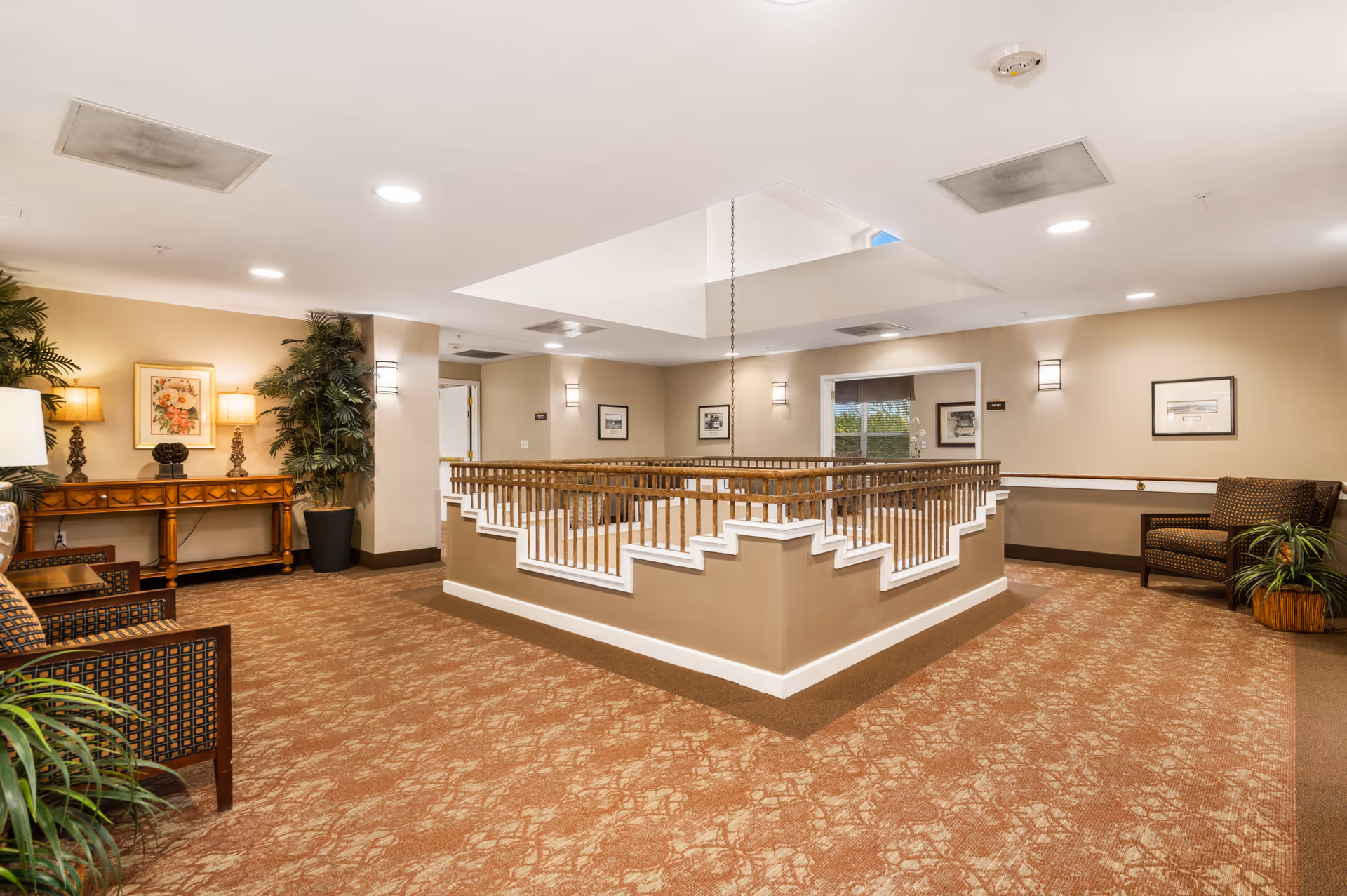 Interior view of a well-lit common area in an assisted living facility featuring beige walls, patterned carpet, wooden railing around a central open space, several chairs, potted plants, framed artwork on the walls, and a wooden console table with lamps and decorative items.