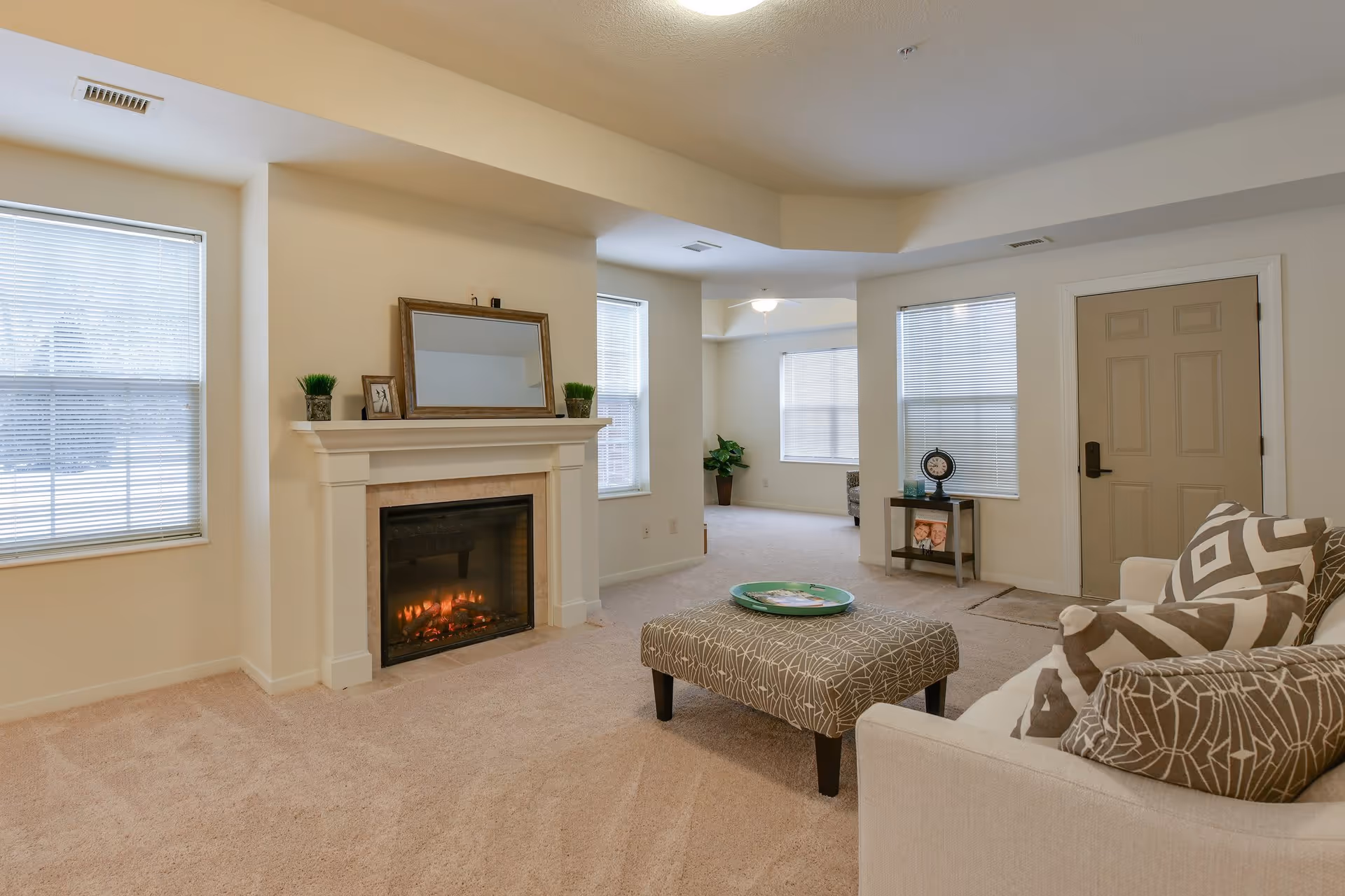 A cozy living room with beige carpet and walls, featuring a white fireplace with a mirror and small plants on the mantel. There is a patterned ottoman with a green tray in front of a beige sofa adorned with patterned pillows. The room has multiple windows with blinds and a beige door, with a small table holding a clock and framed photos near the entrance.