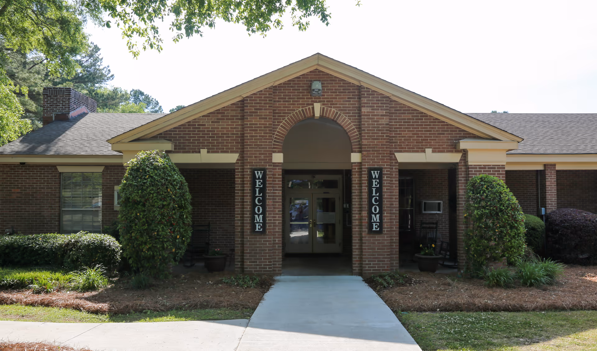 Front exterior view of a single-story brick building with a peaked roof and an entrance walkway. Two vertical signs on either side of the entrance read 'WELCOME'. There are trimmed bushes and landscaping around the entrance.