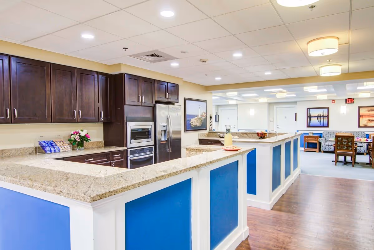 Bright communal kitchen and seating area with granite counters, blue-paneled islands, dark wood cabinets and a lounge visible in the background.