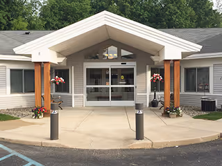 Front entrance of a single-story building with a covered porch supported by wooden columns. There are flower pots with red and white flowers on either side of the entrance, a concrete walkway, and a parking area in front. The building has light-colored siding and several windows.