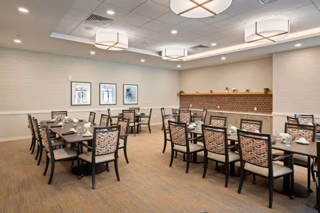 A dining room with several rectangular tables and chairs arranged neatly. Each table is set with white cups, saucers, and small flower arrangements. The chairs have patterned upholstery with a geometric design. The room has beige walls, three framed abstract artworks on one wall, and modern ceiling lights. There is a counter with cabinets and a brick backsplash along one wall.