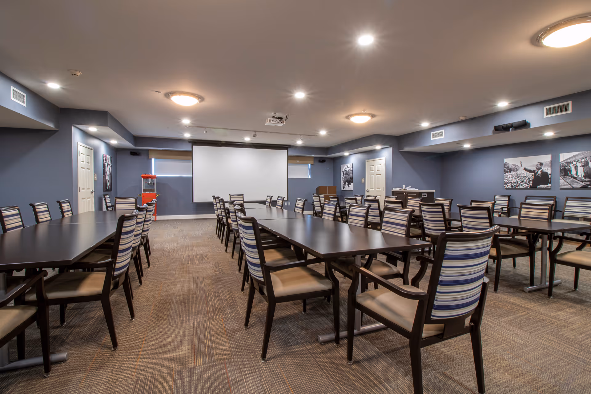 A large meeting or activity room with multiple long tables and striped cushioned chairs arranged in rows. The room has gray walls, carpeted floor, ceiling lights, a projector mounted on the ceiling, and a large white projection screen on one wall. There are black and white framed photos on the walls and a red popcorn machine in the corner.