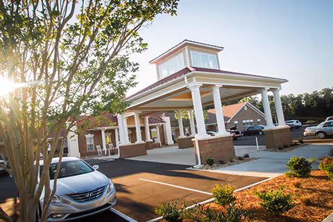 Exterior view of Lake Park Nursing and Rehabilitation Center showing the entrance with a covered drop-off area supported by white columns, a tree in the foreground, parked cars, and a clear sky.
