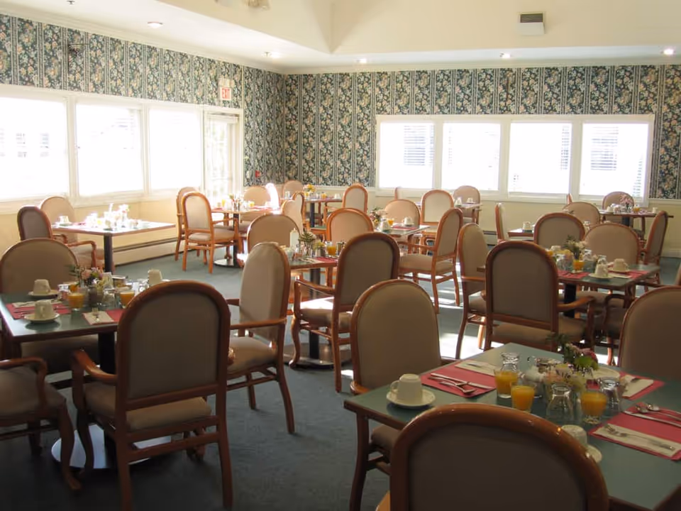 Sunlit dining room with multiple tables and chairs set for breakfast, floral wallpaper, and large windows.