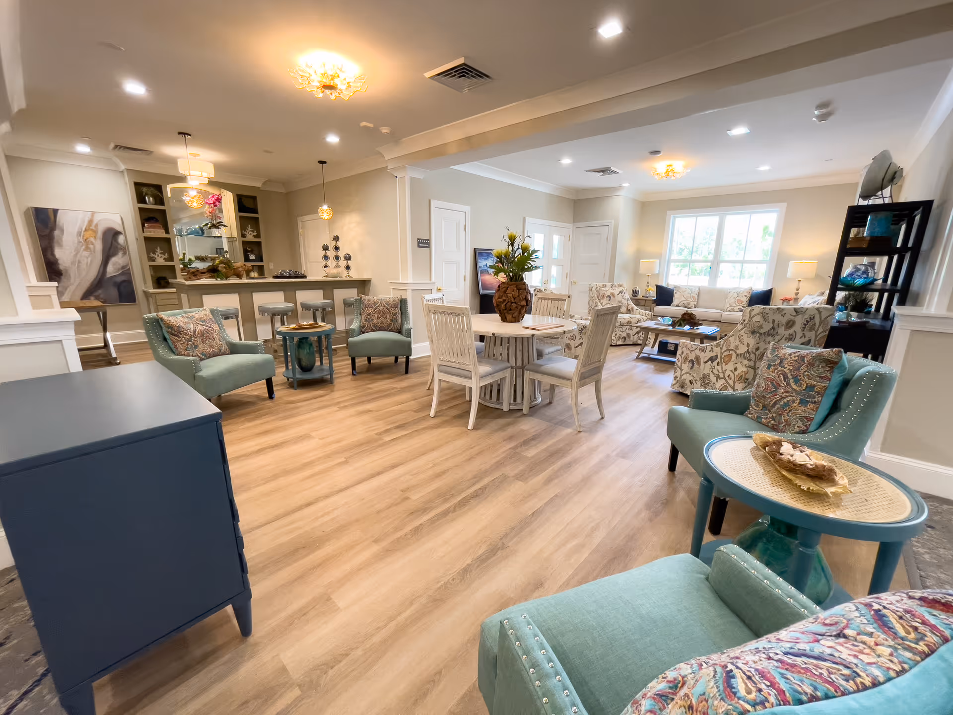A spacious and well-lit senior living common area with light wood flooring, multiple seating options including teal armchairs with patterned cushions, a round wooden table with chairs, and a beige sofa set near a large window. The room features modern ceiling lights, a small bar area with stools, decorative shelves, and tasteful artwork on the walls.