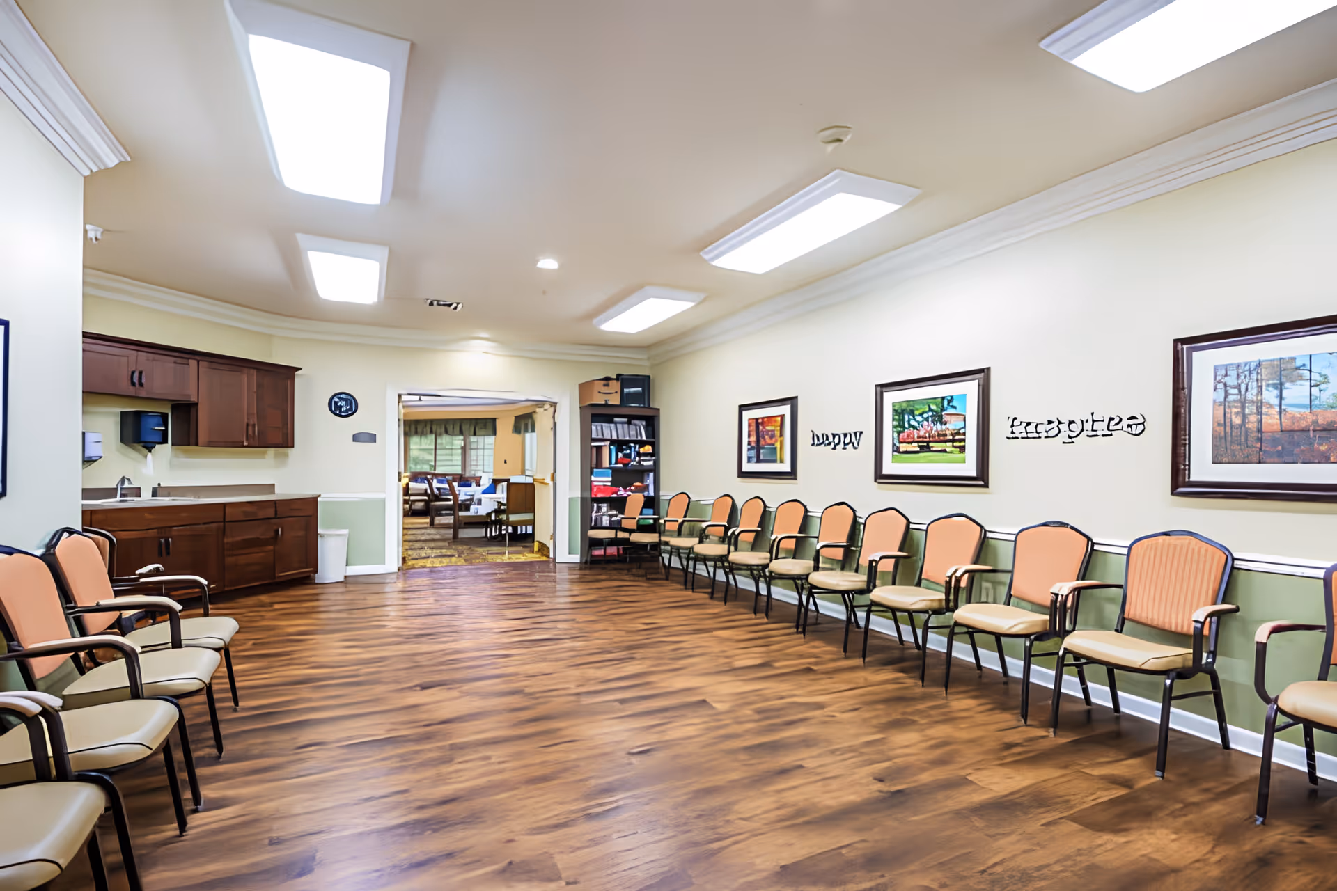 A spacious senior living common room with rows of chairs along the walls, hardwood floors, framed artwork, and a small kitchenette area.