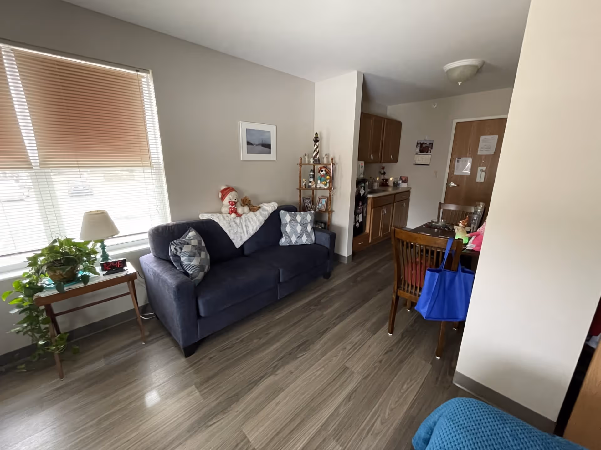 A cozy living area in a senior living facility with a dark blue couch adorned with patterned pillows and stuffed toys. Next to the couch is a small wooden side table with a lamp, a digital clock showing 12:46, and a green potted plant. The room has wood flooring, a window with blinds, a small dining table with chairs, and a kitchenette area with cabinets and a countertop. The walls are light-colored, and there is a framed picture hanging above the couch.