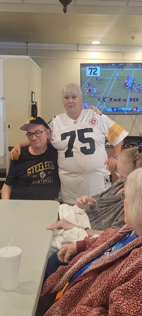 A group of elderly people sitting around a table in a common area. One woman wearing a white football jersey with the number 75 is standing and has her arm around a man wearing a black Steelers football shirt and cap. A television in the background shows a football game.