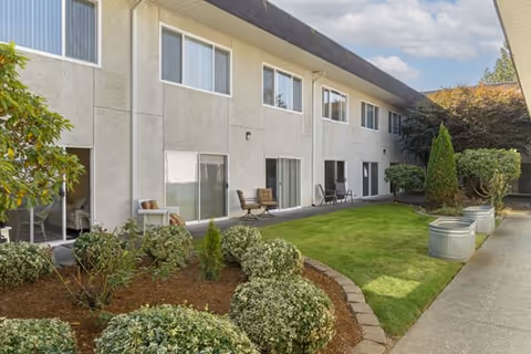 Outdoor view of a senior living facility with a two-story building featuring multiple windows and sliding glass doors. The area includes a well-maintained lawn, landscaped bushes, small trees, and patio chairs placed outside some of the ground floor units under a partly cloudy sky.