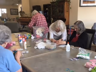 Several elderly women seated around a table engaged in arts and crafts activities in a communal room. The table is covered with various craft supplies including glue, cotton balls, and colorful materials. The room has comfortable seating and cabinets in the background.