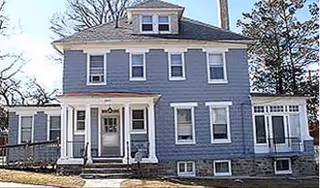 Exterior view of a two-story blue house with white trim, featuring multiple windows, a front porch with steps, and a ramp on the left side. The house is surrounded by trees and has a stone foundation.
