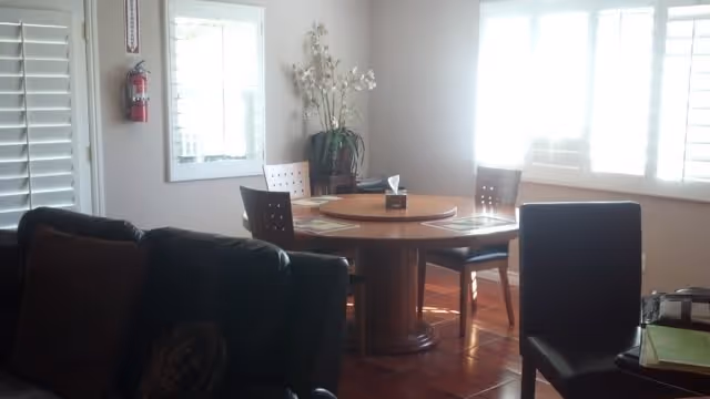 A bright room with a round wooden dining table surrounded by four chairs. There is a large window with white blinds letting in natural light. A dark sofa is partially visible in the foreground, and a tall vase with white flowers is placed near the window.