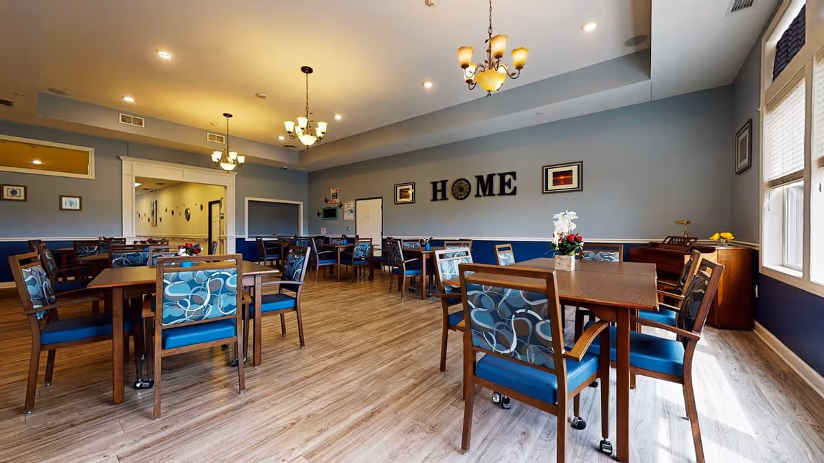 A spacious dining room with multiple wooden tables and chairs featuring blue patterned cushions. The walls are painted light blue with a darker blue wainscoting. There are three chandeliers hanging from the ceiling providing warm lighting. On one wall, the word 'HOME' is displayed in large decorative letters along with framed pictures. Large windows on the right side let in natural light, and there is a wooden sideboard with flowers and decorative items near the windows.