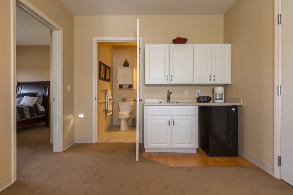 Interior view of a room in an assisted living facility showing a small kitchenette with white cabinets, a sink, a coffee maker, and a mini refrigerator. To the left, there is an open door leading to a bathroom with a toilet, towel racks, and wall decorations. Further left, there is a glimpse of a bedroom with a bed and pillows.