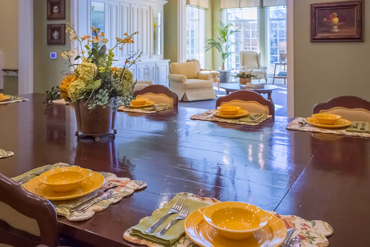 A dining table set with yellow plates and bowls, silverware, and floral placemats. A large floral centerpiece is in the middle of the polished wooden table. In the background, there is a living area with armchairs, a coffee table, and large windows letting in natural light.