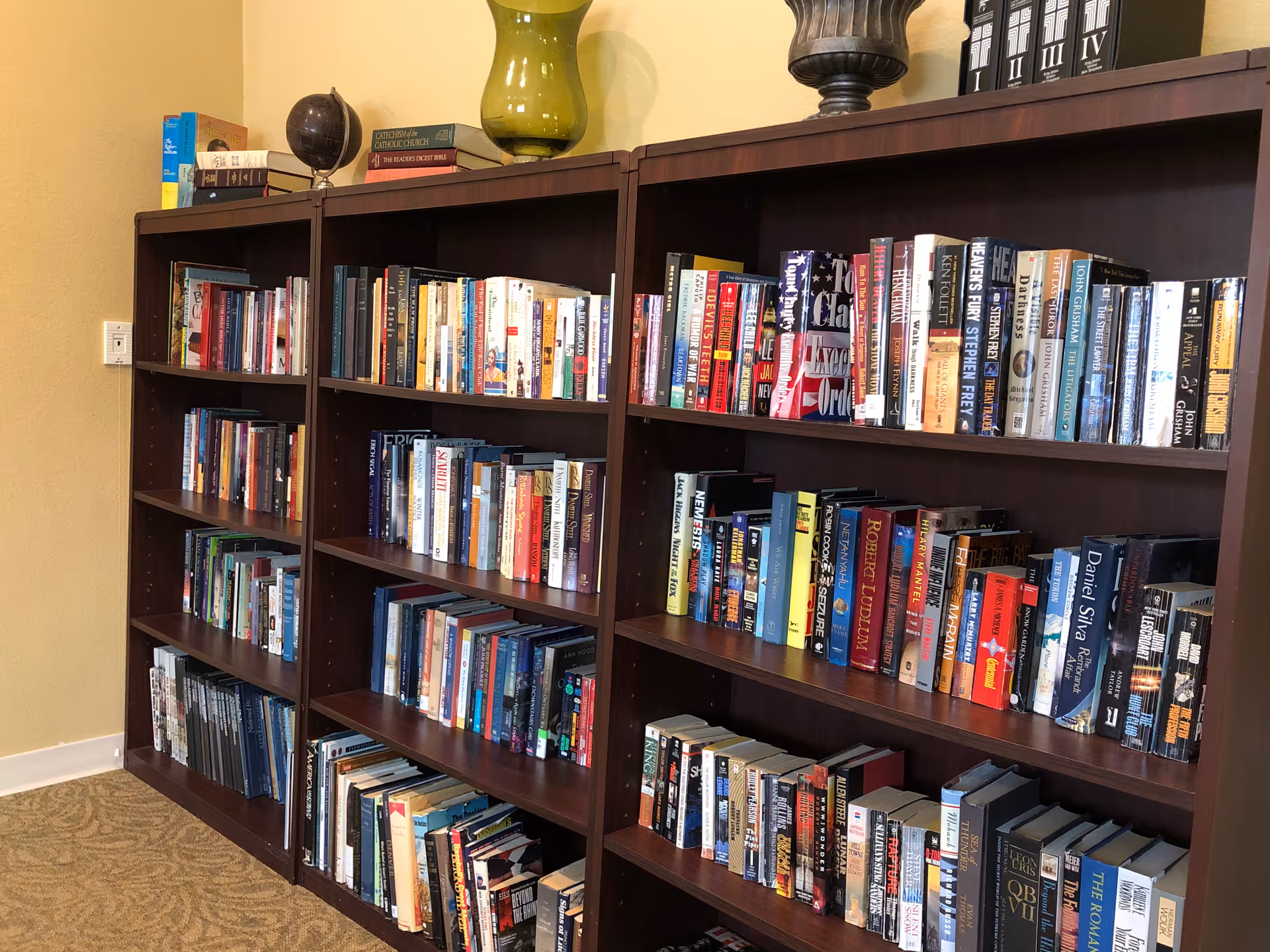 A wooden bookshelf filled with a variety of books in a room with beige walls and carpeted floor. On top of the bookshelf are decorative items including a green vase, a globe, and some black and white binders.
