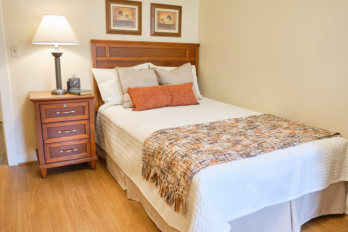 A neatly made bed with white bedding, several pillows including a long orange one, and a patterned throw blanket at the foot. Next to the bed is a wooden nightstand with three drawers, a table lamp, a clock, and a book. Two framed pictures hang on the wall above the headboard. The room has light-colored walls and wooden flooring.
